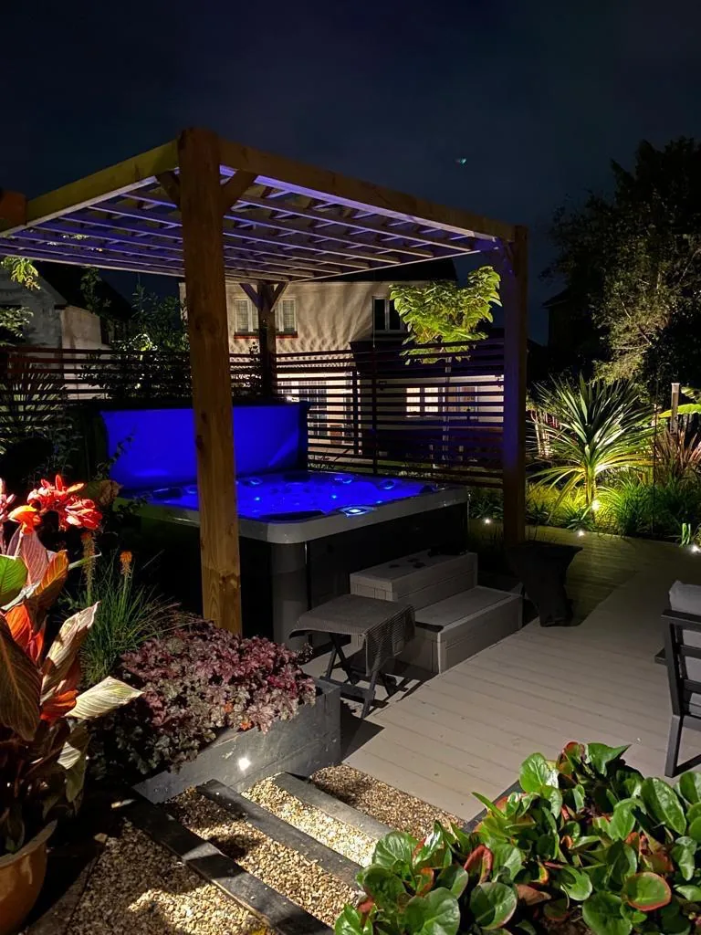 Lit pergola at dusk with festoon lights over composite decking and tropical planting