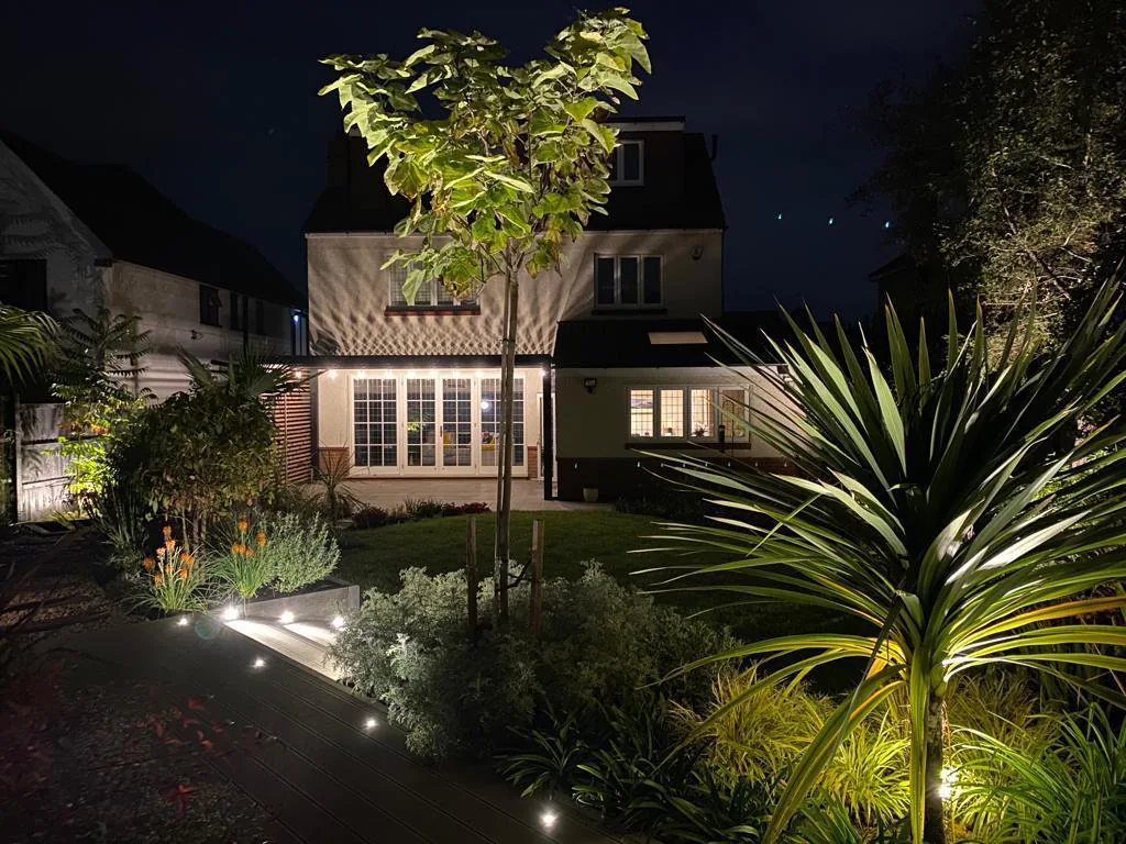 Front garden at night with the house warmly lit and mature ornamental planting in uplighting
